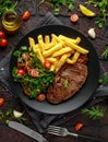 Grilled sirloin steak with potato fries and vegetables, tomato salad in a black plate. rustic table Royalty Free Stock Photo