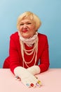 Grimace face. Beautiful old woman, grandmother in stylish red dress and pearl necklace posing over blue studio Royalty Free Stock Photo