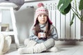Grimacing little girl in hat, socks, pullover drink sour medicine syrup and sit near heating convector. Home treatment