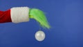 The Grinch's green haired hand holds a white Christmas ball on an isolated blue background. Gift kidnapper cosplay Royalty Free Stock Photo