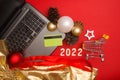 Grocery cart, laptop with bank credit card, christmas balls and pine cones and number 2022 on red background. Christmas online Royalty Free Stock Photo