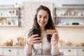 Grocery shopping app. Cheerful woman using credit card and cellphone, buying food online while sitting in kitchen Royalty Free Stock Photo