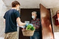 A grocery store delivery man wearing a black polo-shirt delivering food to an old man at home