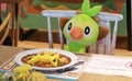 Grookey plushie sitting at a table in the PokÃÂ©mon CafÃÂ© with plastic food on it.