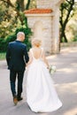 Groom in a black suit and bride in a white dress with a bouquet of flowers are walking holding hands along the road to Royalty Free Stock Photo