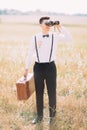 The groom in black suit decorated with air-balloon is carrying the vintage suitcase and looking through binoculars at Royalty Free Stock Photo