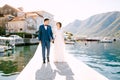 Groom and bride walk along the pier holding hands against the background of the buildings. Mountains and floating boats Royalty Free Stock Photo