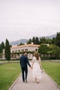 Groom and bride in a white dress, holding hands, walk along the path among the garden to the old villa among the green Royalty Free Stock Photo