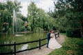 The groom in a dark suit hugs the girl in a lush white dress. Lovers walk in the park with a pond and a fountain.