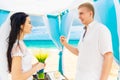Groom giving an engagement ring to his bride under the arch decorated with flowers on the sandy beach. Wedding ceremony on