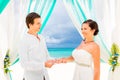 Groom giving an engagement ring to his bride under the arch decorated with flowers on the sandy beach. Wedding ceremony on