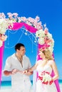 Groom giving an engagement ring to his bride under the arch decorated with flowers on the sandy beach. Wedding ceremony on Royalty Free Stock Photo