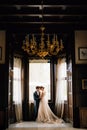 Groom hugs bride against the background of the large windows of the old villa. View from the next room with a chandelier Royalty Free Stock Photo