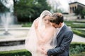 Groom hugs bride in a veil from behind and kisses her on the shoulder near the fountain Royalty Free Stock Photo