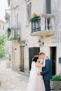 Groom hugs bride in a white dress near a stone building with balconies