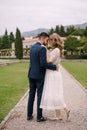 Groom hugs bride in a white dress while standing on a path among the garden on the background of an old villa and green Royalty Free Stock Photo