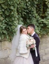 Groom hugs tender bride in white fur coat under the wall