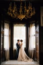 Groom almost kisses bride against the background of the large windows of the old villa. View from the next room with a