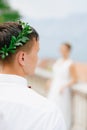 The groom in an olive wreath and a white shirt, in front of a silhouette of the bride in defocus