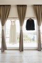 The groom`s suit is black and the bride`s white lace dress on hangers by the window with gray curtains. Royalty Free Stock Photo