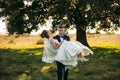 Groom take the bride in his arms and carried her to the field in background of big tree. Groom kisses bride