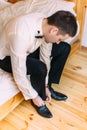 The groom is tying the wedding shoes while sitting on the bed.