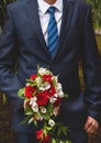 The groom in a white shirt, blue suit and tie holds the wedding bouquet of the bride of white and red flowers of roses Royalty Free Stock Photo