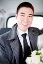 The groom in a winter coat sitting in the car with wedding bouquet in hands. Positive portrait, looking into the camera.