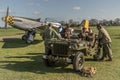 Ground crew sit in a WW2 jeep with a P-51 Mustang in the background.