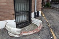 Ground floor window covered in iron grate with crumbling window well and asphalt