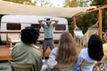 Group of diverse millennial friends playing charades near trailer, trying to guess word on autumn camping vacation