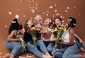 Group of diverse women with bouquets having fun under falling petals. Six happy females sitting together against a brown