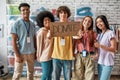 Group of diverse young volunteers smiling at camera while standing in charitable organization office, Guy holding card