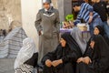 A group of elderly Muslim women wearing black burqas are sitting on the sidewalk by the bazaar