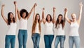 Group of five beautiful diverse young women wearing white shirt and denim jeans raising their arms, looking cheerful at