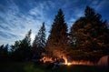 Group of friends sitting around a bonfire on a green mountain meadow surrounded by spruce trees under starry sky Royalty Free Stock Photo