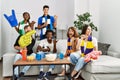 Group of friends supporting football team watching tv al home sitting on the sofa pointing thumb up to the side smiling happy with