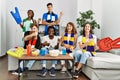 Group of friends supporting football team watching tv al home sitting on the sofa smiling happy pointing with hand and finger to