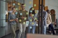 Group of happy creative people or coworkers looking at sticky notes on glass board and smiling, discussing new project