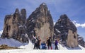 A group of joyful hikers pose happily and proudly at the base of three huge mountain walls with snow and beautiful blue skies. Tre