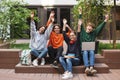 Group of joyful students sitting and happily looking in camera while raising their hands up in courtyard of university