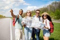 Group of multiethnic friends checking map, choosing route during road trip, hitchhiking on highway