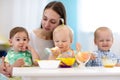Group of nursery babies eat healthy food with kindergartener help. Lunch break in creche. Little children have dinner in