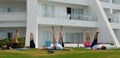 Group of people doing Yoga on grass outside hotel with legs in the air.