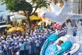 People marching in a parade, with blue traditional clothes and a large white artificial bird above