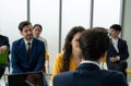 A group of people are sitting in a room, some of them wearing suits. A man in a suit is talking to a woman in a yellow dress. Royalty Free Stock Photo