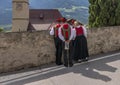 A group of people in traditional Tyrolean clothing looks out from a low wall to look towards the parish church of Corzes, Italy