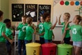 Group of schoolchildren holding color coded recycling bins and bags in an elementary school classro