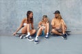 Group Of Skaters With Skateboards. Hipsters In Casual Outfit Sitting Against Concrete Wall At Skatepark