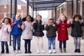 A group of smiling young multi-ethnic school kids wearing coats and carrying schoolbags standing in a row in walkway outside their Royalty Free Stock Photo
